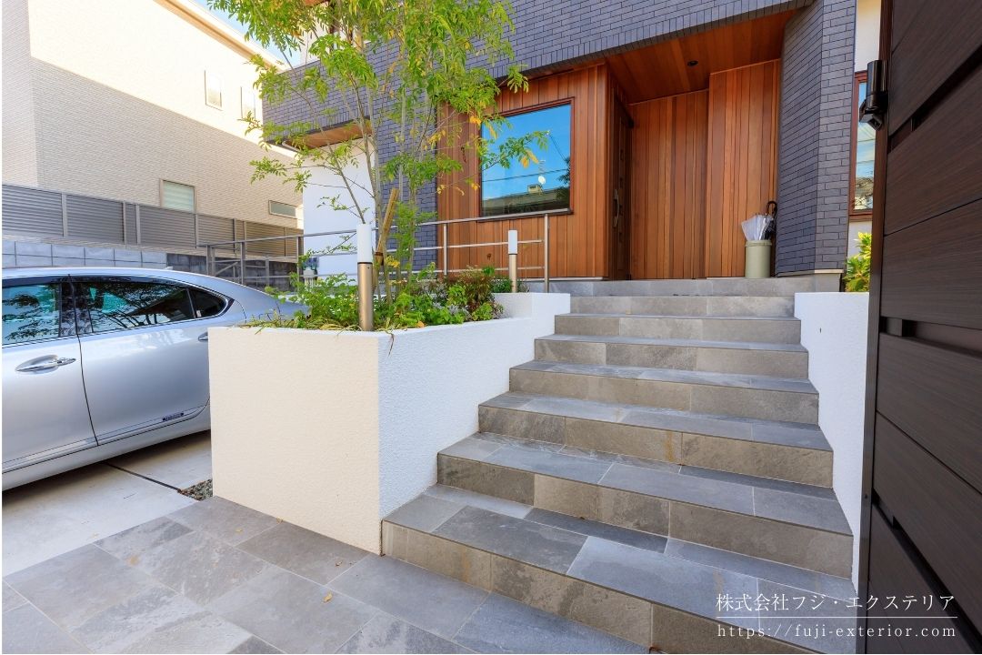 門扉を抜けると、玄関へと続くアプローチ階段があります。その隣には植え込みがあり、シンボルツリーのシマトネリコが爽やかに葉を揺らしています。それはちょど玄関ドアの目隠しにもなっています。45cm幅の広いアプローチには、グレーのタイル、リビエラの フィオラクォーツを貼り、シックな雰囲気を醸し出しています。